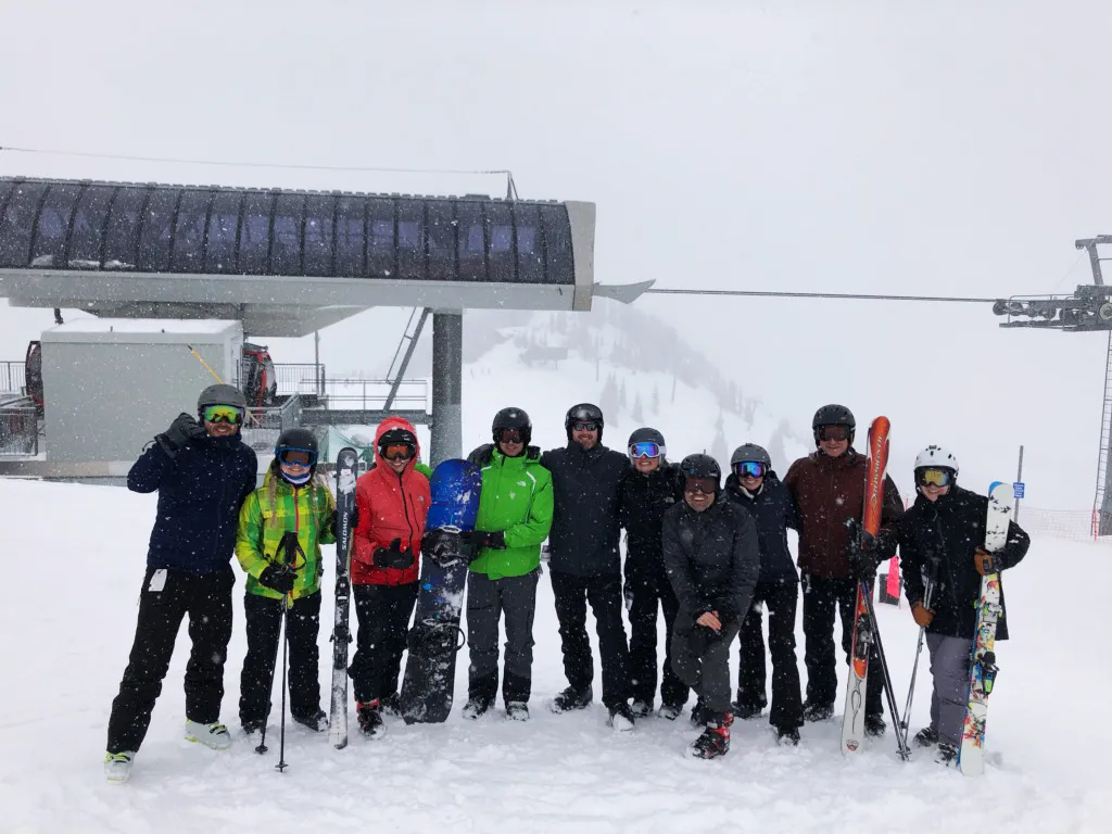 Group photo in front of ski lift, including people with snowboards and skis